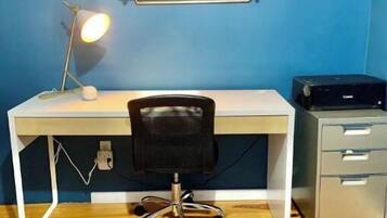 Ein Zimmer mit weißem Schreibtisch, schwarzem Bürostuhl, gestreiftem Teppich, einer Lampe, einem Regal mit einer Fuchsfigur und einem Drucker.