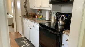 Cocina compacta con armarios blancos, estufa negra y microondas. Un tazón de madera y un racimo de plátanos están sobre la encimera.