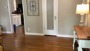 Una habitación con piso de madera, una puerta blanca, un reloj de pared y una mesa con una lámpara.