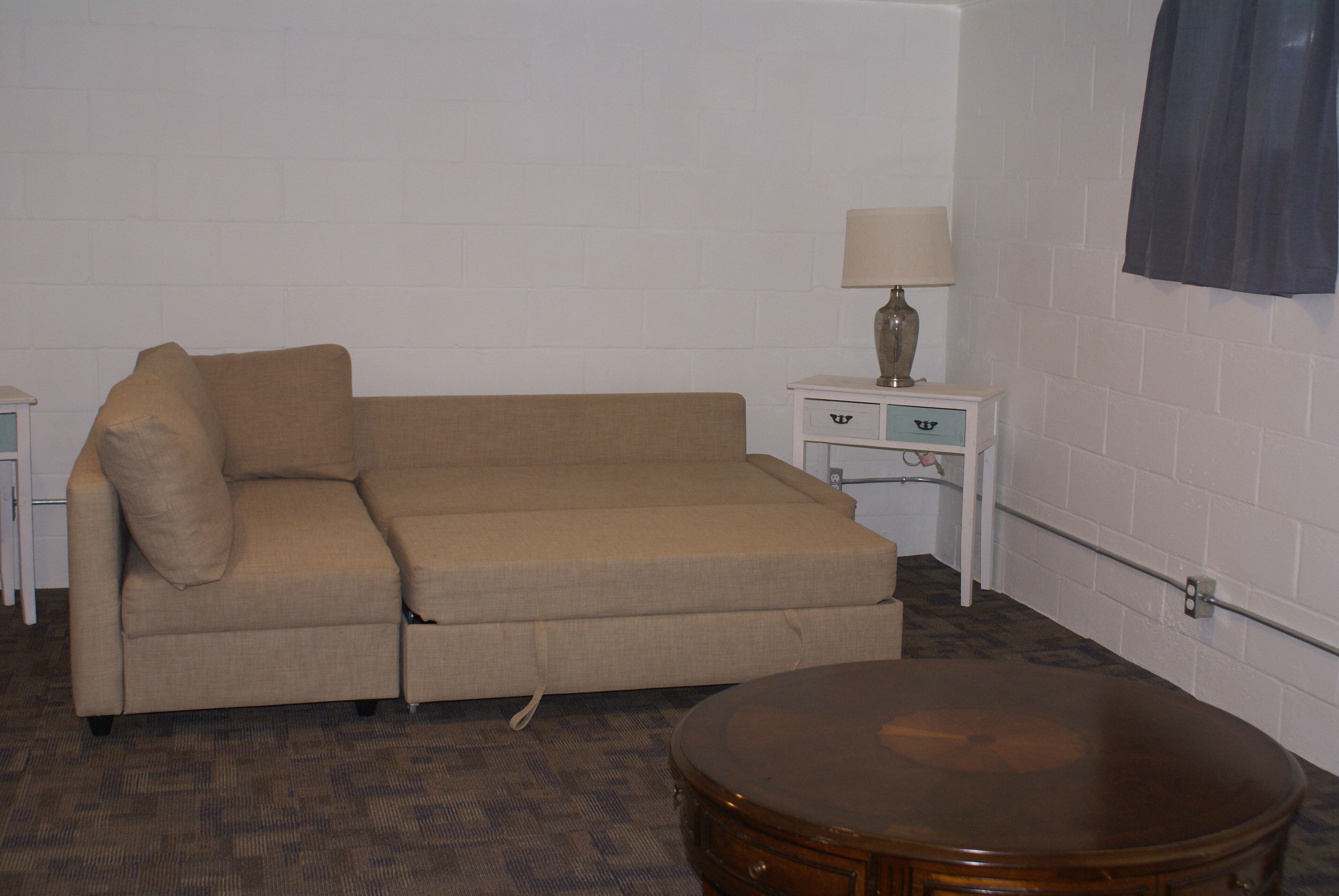 A beige sofa in a room with a patterned carpet, a small wooden table, and a nightstand with a lamp.