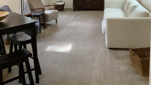 Una sala de estar con ventilador de techo, televisor de pantalla plana montado en la pared, sofá blanco, mesa de comedor con sillas y un ventanal con persianas.