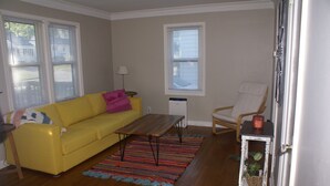 Una sala de estar con un sofá amarillo, una mesa de centro de madera, un tapete colorido, una silla blanca y una mesita con una planta.