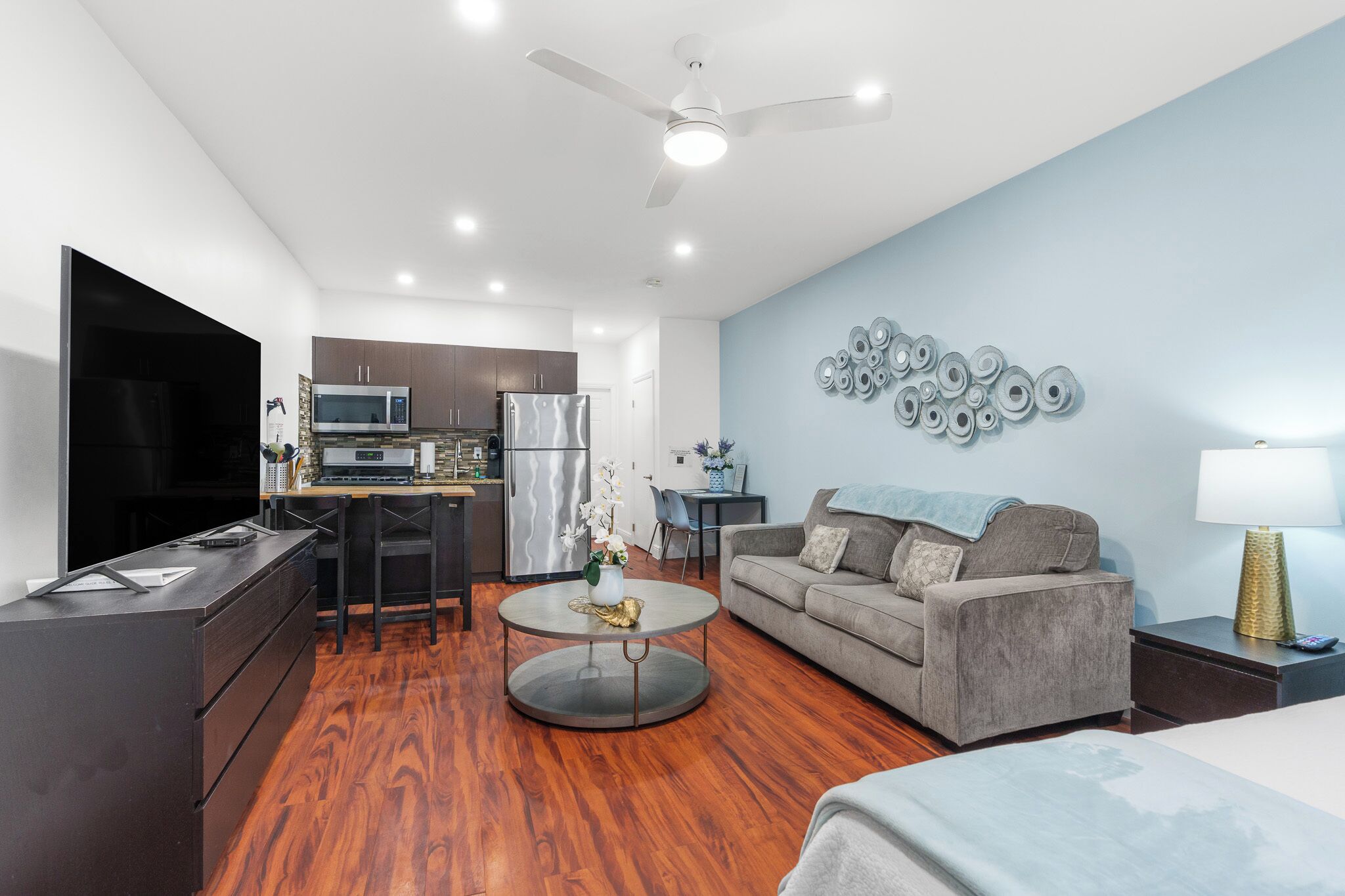 A modern living room with a flat-screen TV, a sofa, a coffee table, and a dining area with a microwave and refrigerator.