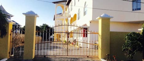 A gated entrance to a building with a white facade and yellow accents, featuring a central archway and a cobblestone driveway.