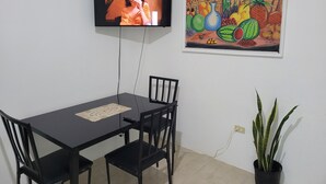 Ein Essbereich mit an der Wand montiertem Fernseher, einem gerahmten Gemälde, einer Topfpflanze und einer Fernbedienung auf dem Tisch.