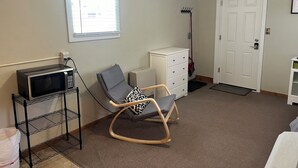 Ein Zimmer mit Schaukelstuhl, weißem Schminktisch, eingebauter Mikrowelle, einer Tür und einem Fenster mit Jalousien.