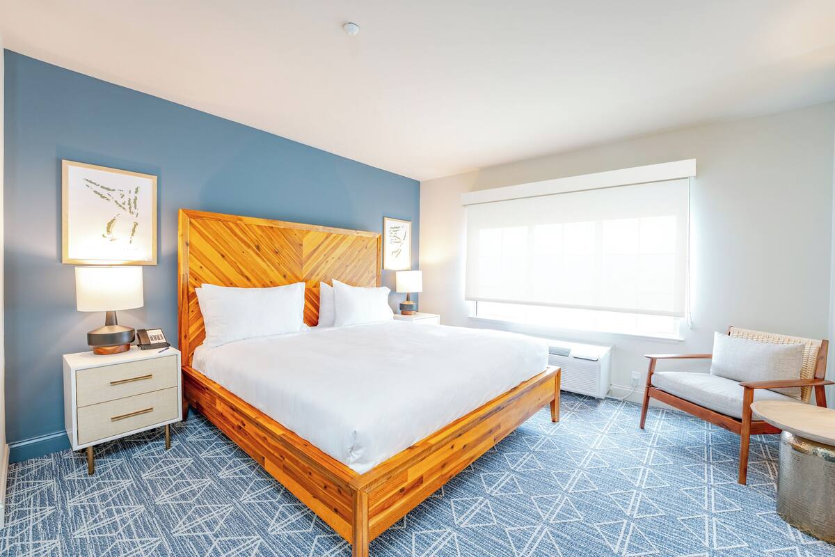 A hotel room with a wooden bed, a nightstand with a lamp, a chair, and a large window.