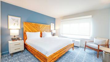 Quarto de hotel com cama de madeira, mesa de cabeceira com abajur, uma cadeira e uma janela grande.