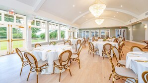A spacious banquet hall with round tables and wicker chairs, large windows, and a high ceiling with a chandelier.