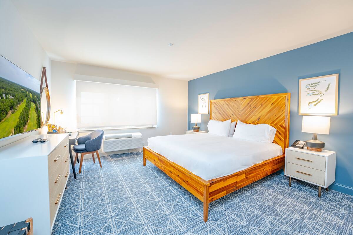 A bedroom with a large bed, a desk, and a chair. There is a window with a view of greenery.