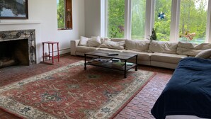 Uma sala de estar com lareira, sofá, mesa de centro e grandes janelas com vista para o verde.
