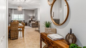 A modern living room with a dining area, featuring a wooden dining table, wicker chairs, a sofa, and a coffee table. There is a decorative mirror on the wall and a sign that reads "WELCOME TO PALMS OF PARADISE".