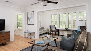 Ein Wohnzimmer mit Sofa, Couchtisch und Fernseher. Ein Fenster mit Jalousien, ein Deckenventilator und eine Pflanze auf dem Couchtisch.