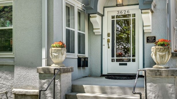 Ein Haus mit einer weißen Tür, auf der „3626“ steht, einer Veranda mit Stufen und zwei großen, blumengeschmückten Pflanzgefäßen.