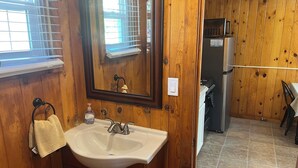 Une salle de bain aux murs lambrissés, équipée d’un lavabo, d’un miroir, d’un porte-serviettes et d’une fenêtre avec des stores.