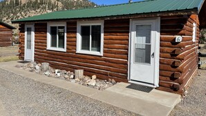 Une cabane en rondins surmontée d’un toit vert, avec des fenêtres à cadre blanc et une porte portant le numéro 6.