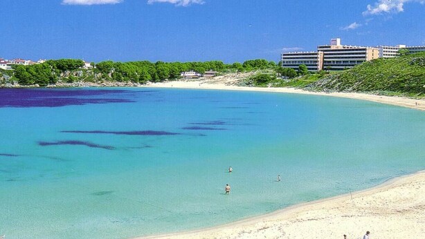 A beach with clear turquoise water, a sandy shore, and a building in the background.