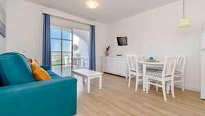 A modern living room with a teal sofa, a dining area with white chairs, and a kitchen with a TV.