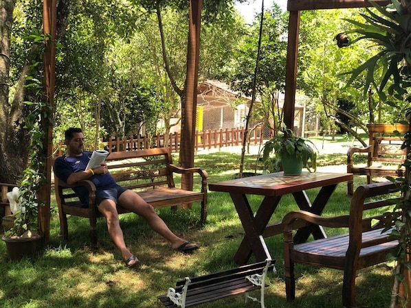A man sitting on a wooden bench in a garden area with a table and chairs nearby.