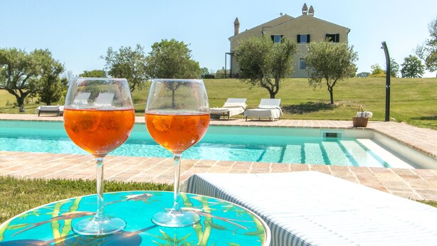 Dos vasos de bebida naranja sobre una mesa junto a una piscina, con una casa al fondo.