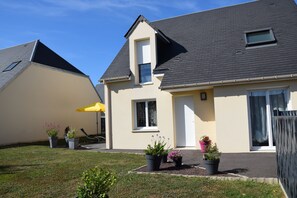 Une maison à deux étages avec un toit en pente, une petite fenêtre et une porte. Des plantes en pot sont placées devant la maison.