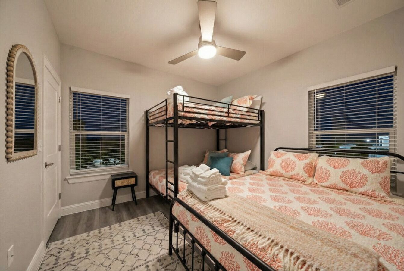 Dormitorio con literas, ventilador de techo, dos ventanas con persianas, un pequeño banco y una alfombra estampada.