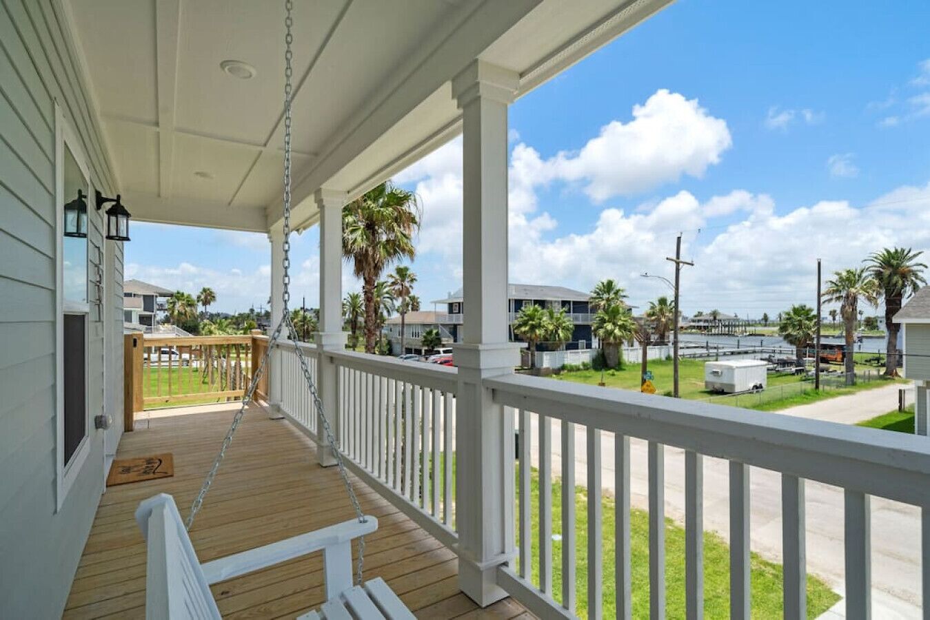 A covered porch with a wooden deck, white railings, and a view of a marina with boats and palm trees.