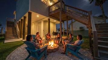 A group of people sitting around a fire pit on a patio with a wooden deck and stairs leading to a house.