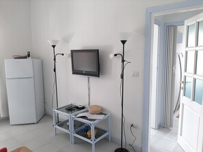 Un espace de vie compact comprenant un réfrigérateur blanc, une petite table avec un téléviseur fixé au mur et un grand lampadaire.