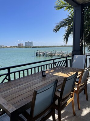 Een houten tafel met stoelen op een balkon met uitzicht op een jachthaven, met boten en gebouwen op de achtergrond.
