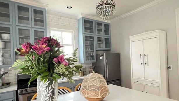 Une cuisine moderne avec des meubles bleu clair, un îlot central blanc, un lustre et un réfrigérateur.