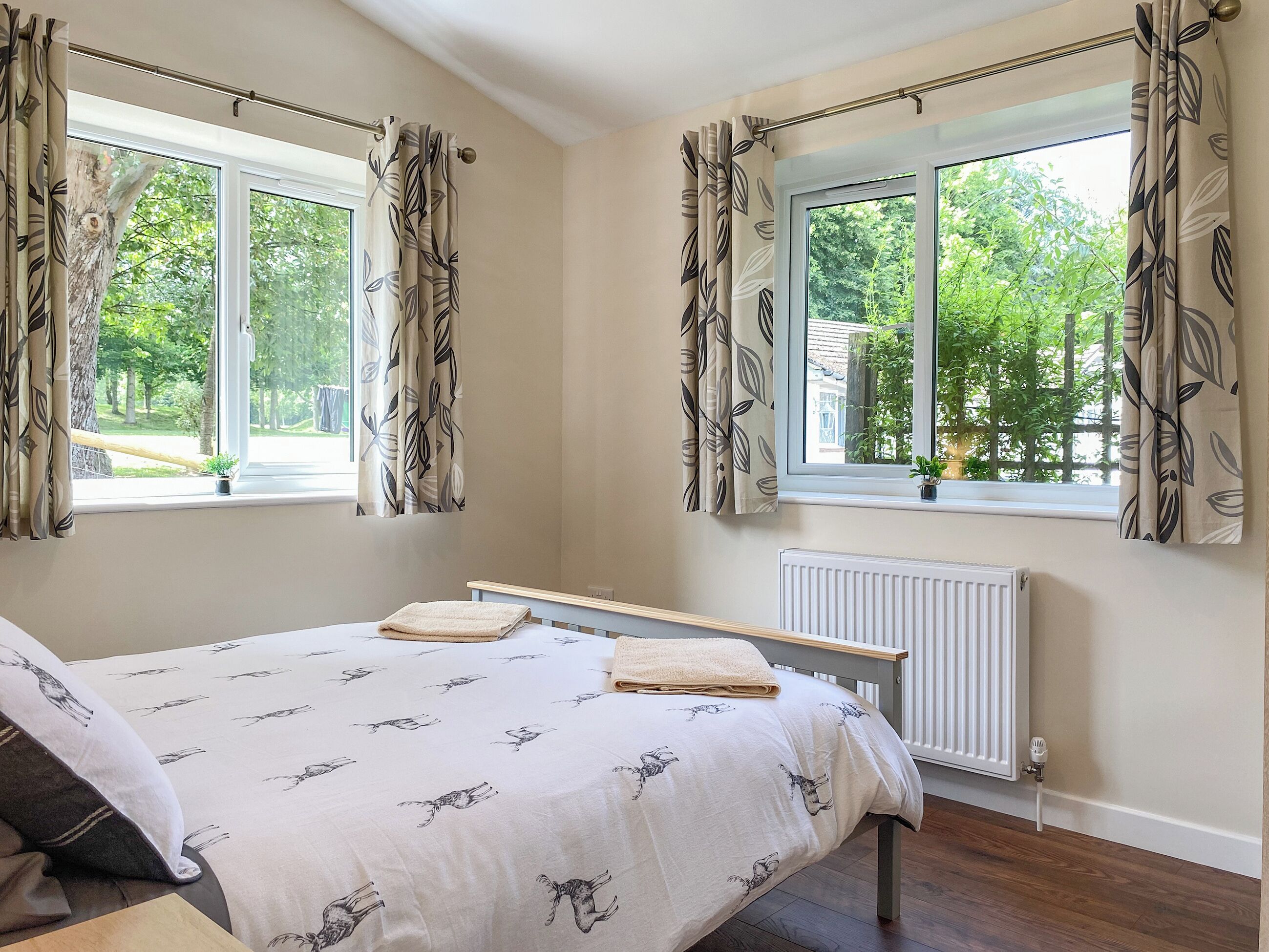 Une chambre à coucher avec un lit, deux fenêtres avec des rideaux, un radiateur et une vue sur la verdure à l’extérieur.
