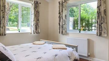 Une chambre à coucher avec un lit, deux fenêtres avec des rideaux, un radiateur et une vue sur la verdure à l’extérieur.
