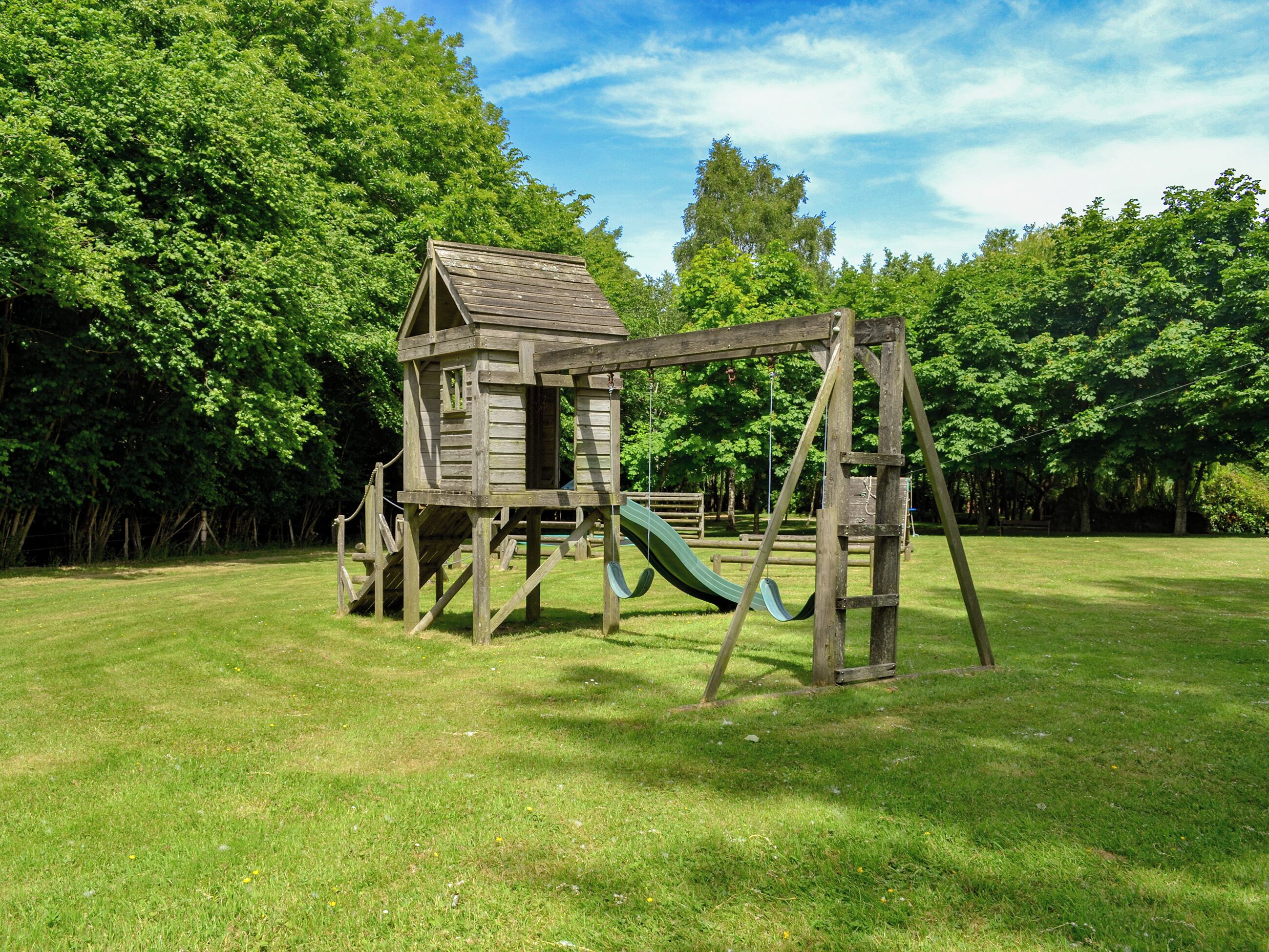 A wooden playhouse with a slide and swings in a grassy park area.