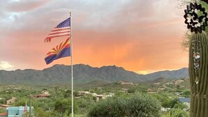 Un asta con la bandera estadounidense y la bandera del estado de Arizona, rodeada de cactus y plantas en macetas, con montañas al fondo.