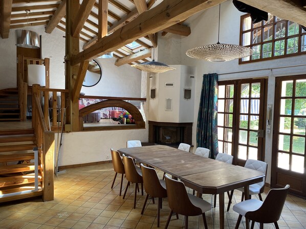 Uma sala de jantar ampla com uma longa mesa de madeira e cadeiras, lareira e grandes janelas com cortinas.