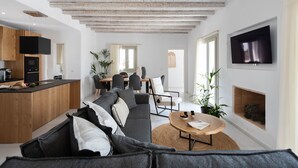 Un salon moderne avec un plafond en bois, un téléviseur noir fixé au mur, un canapé avec des coussins, un coin repas avec des chaises et un coin cuisine avec un comptoir et des appareils électroménagers.