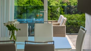 Een balkon met een glazen tafel, witte stoelen en een rieten stoel met kussens. Er staan twee glaasjes op de tafel en een potplant is te zien.