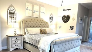 A bedroom with a tufted headboard, a bedside table with drawers, a patterned bedspread, and wall art including a heart-shaped decoration and inspirational phrases. - Quiet peaceful Stone mountain fun House with Pool ,Hot Tub￼ and vineyards near  (Traphill)
