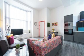 A modern living room with a sofa, a coffee table, and a TV. There is a kitchen area with a microwave and oven.
