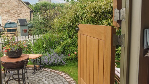A garden with a wooden gate, a brick building, and a tower in the background. - This cosy and quiet Shepherd’s Hut is a great way to unwind. (Frampton-on-Severn)
