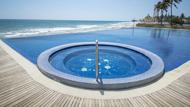 A circular infinity pool with a hot tub, surrounded by a wooden deck, overlooking a beach with palm trees and a clear sky.