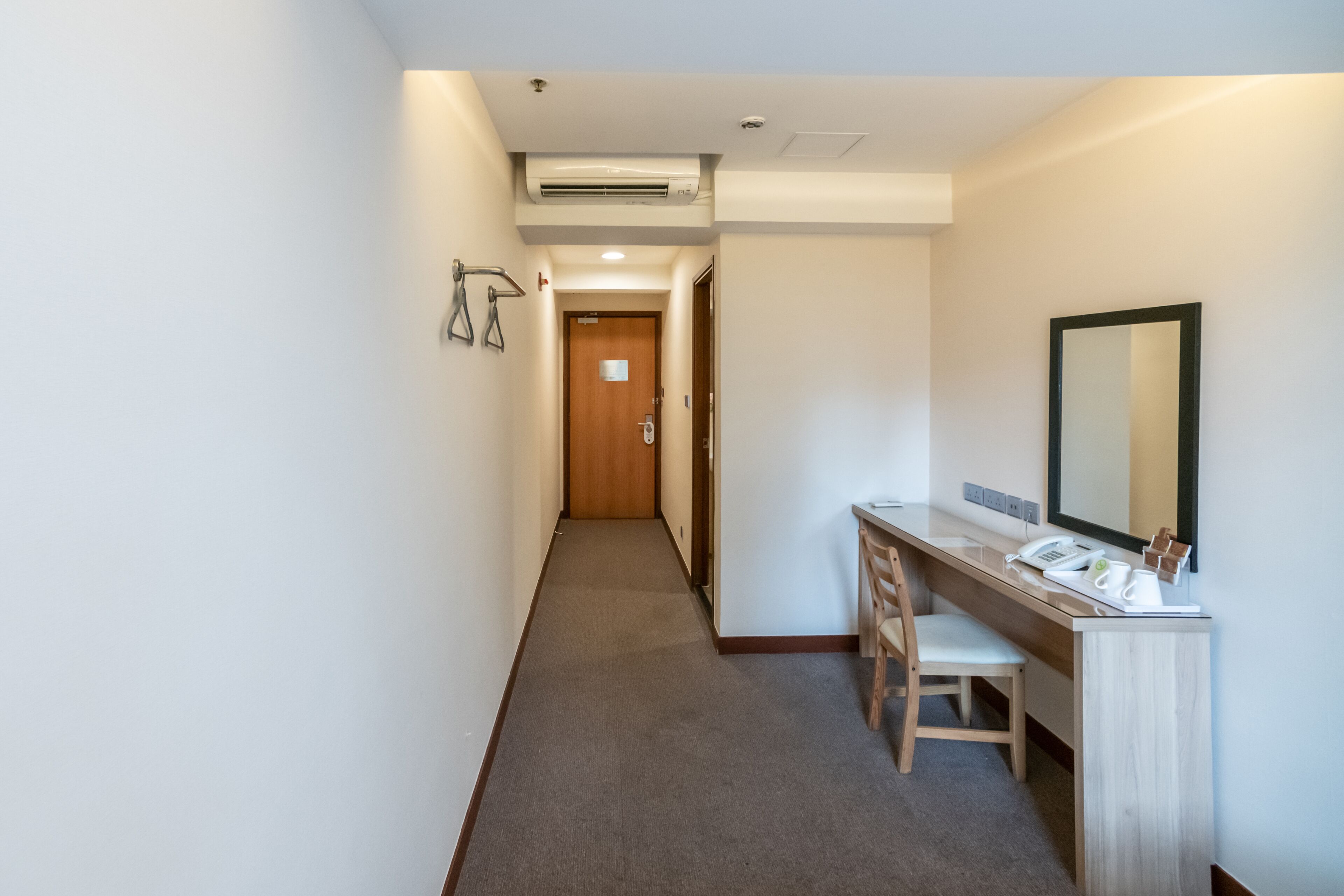 A hotel hallway with a wooden door at the end, a desk with a mirror, and a telephone.