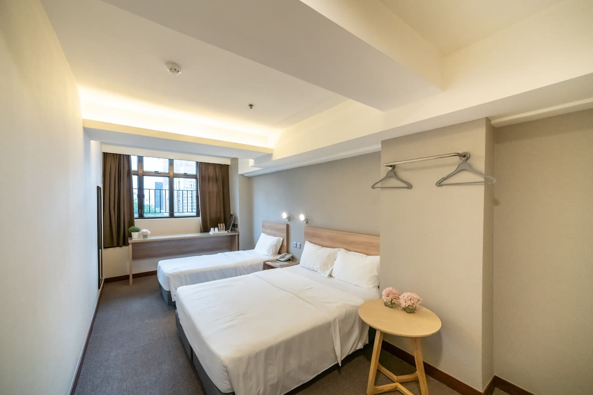 A hotel room with two beds, a bedside table with a lamp, a small table with flowers, and a window with curtains.