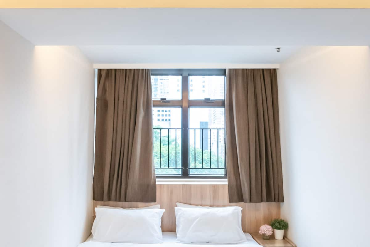 A neatly made bed with white linens, a wooden nightstand with a small potted plant, and a window with curtains.