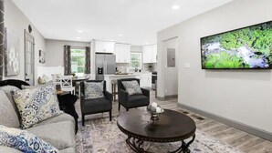 A modern living room with a gray sofa, two black armchairs, and a round coffee table. A kitchen is visible in the background with white cabinets and a large TV mounted on the wall.