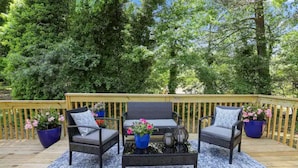 A wooden deck with outdoor furniture, including two chairs and a table, surrounded by potted plants and a blue rug.