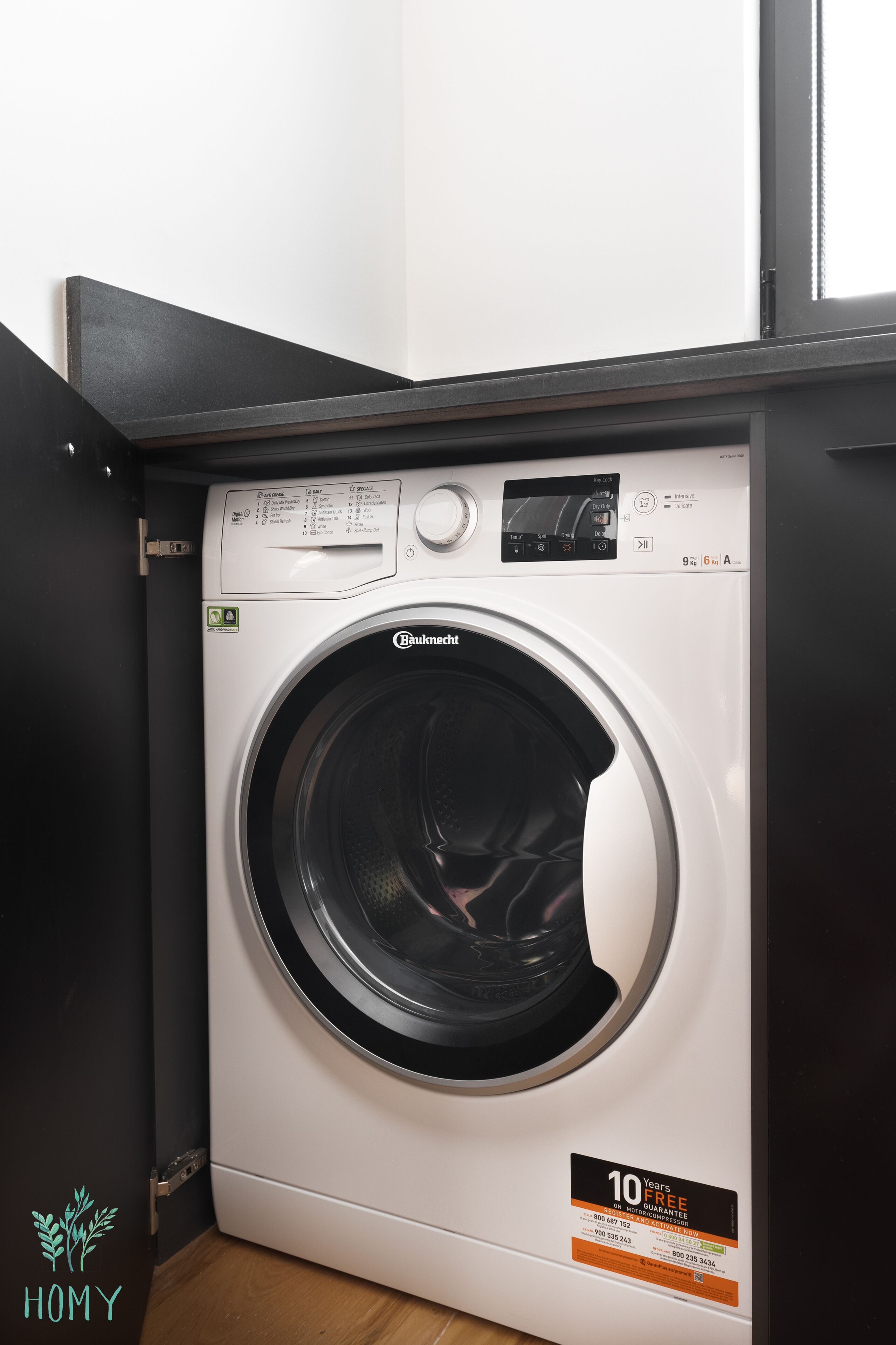 A front view of a Bauknecht front-loading washing machine with a 10-year free guarantee sticker, located in a room with a wooden floor and a black countertop.