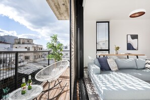 A modern living room with a sofa, a dining table, and a balcony with white chairs and a potted plant. - CARMEL MARKET - Exclusive Collection - By HOMY (Tel Aviv)