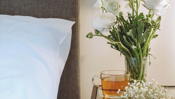 A neatly made bed with white sheets, a bedside table with a glass vase of white flowers, and a glass of amber liquid. - CARMEL MARKET - Exclusive Collection - By HOMY (Tel Aviv)